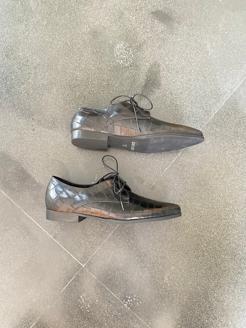 May include: A pair of black leather lace-up shoes with a crocodile-embossed pattern. The shoes have a pointed toe and a low heel.
