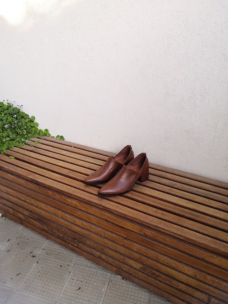 May include: A pair of brown leather heeled shoes with pointed toes are sitting on a wooden bench.
