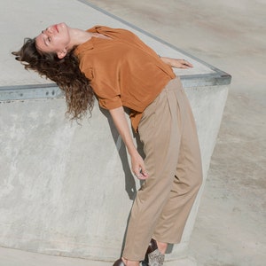 May include: A woman wearing a rust-colored shirt and beige pants is lying on a concrete surface. She is wearing brown and white patterned shoes.