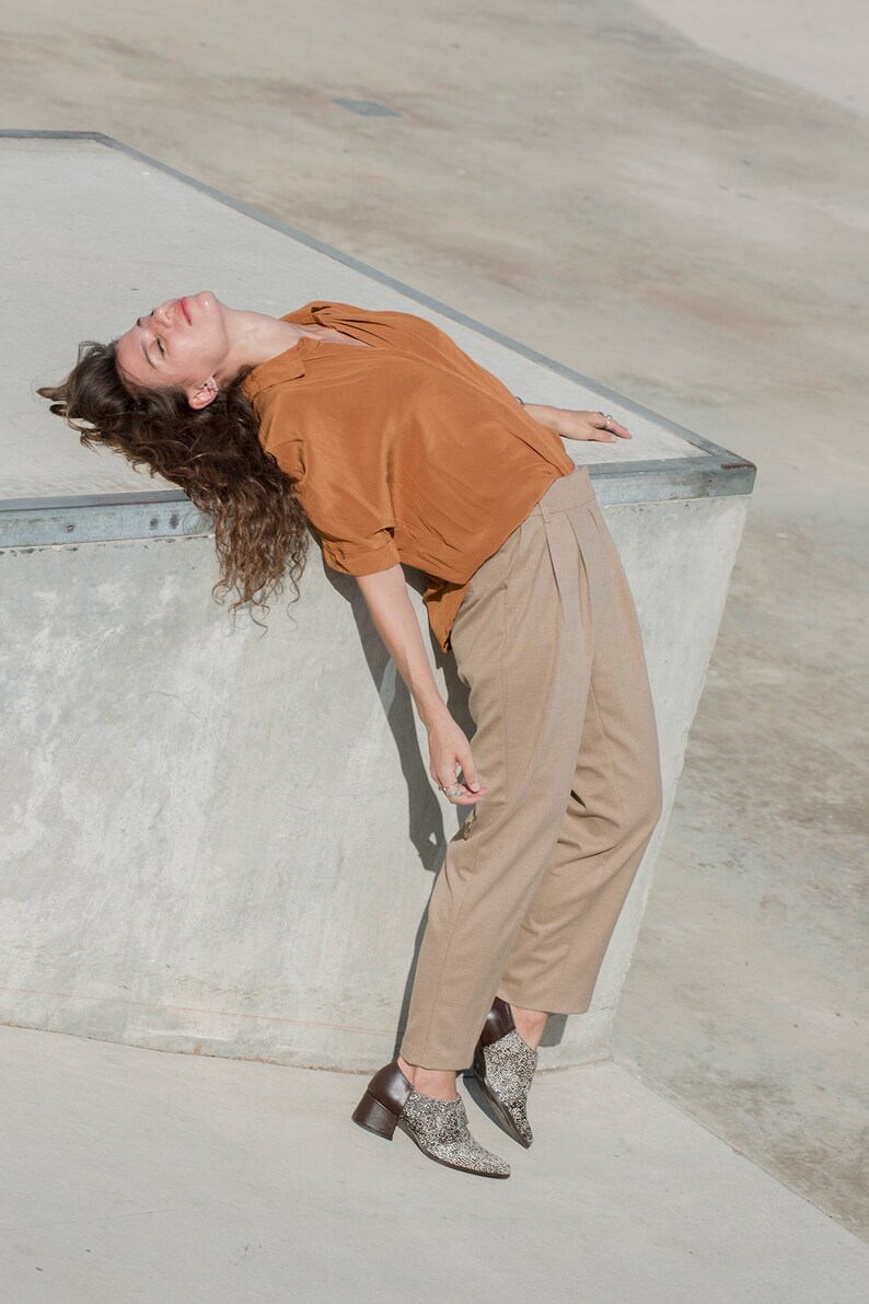 May include: A woman wearing a rust-colored shirt and beige pants is lying on a concrete surface. She is wearing brown and white patterned shoes.