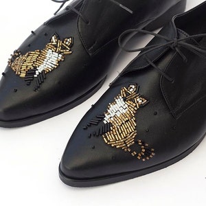 Puede incluir: Zapatos negros de cuero con cordones y adornos de cuentas doradas, blancas y negras en la punta.