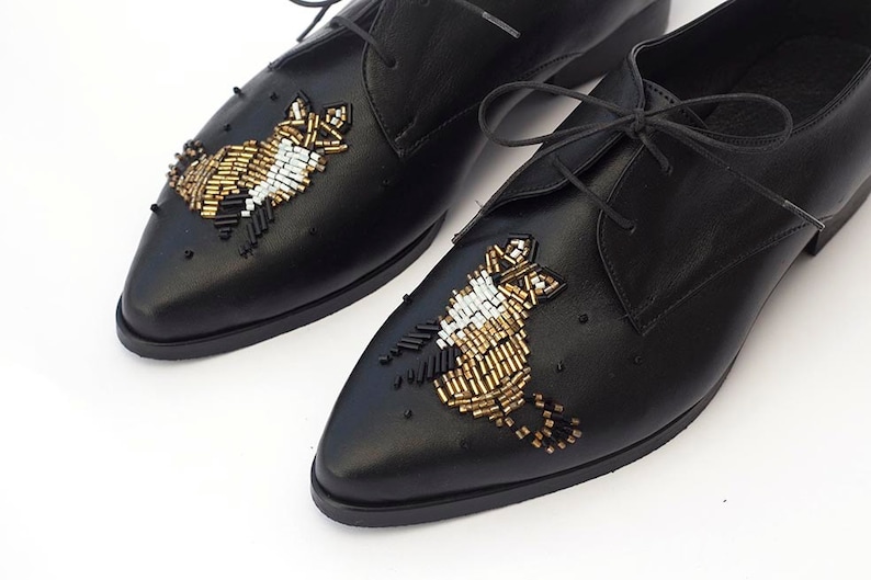 Puede incluir: Zapatos negros de cuero con cordones y adornos de cuentas doradas, blancas y negras en la punta.