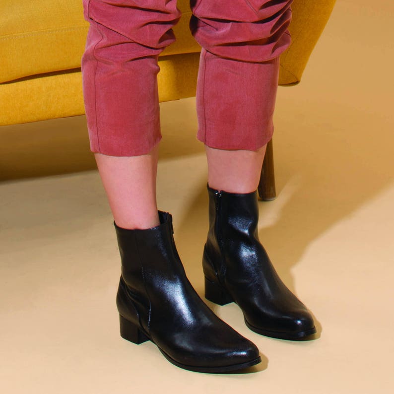 May include: A pair of black leather ankle boots with a pointed toe and a low heel. The boots are worn by a person wearing red pants.