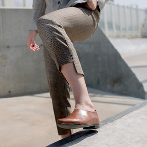 May include: A person wearing brown and tan plaid pants and brown leather shoes. The person is standing on a concrete surface.
