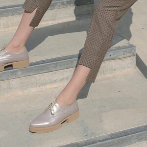 May include: A pair of light gray leather lace-up shoes with a platform sole. The shoes are worn by a person sitting on a set of concrete stairs.