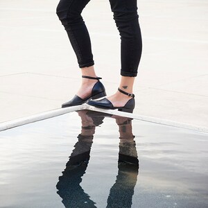 May include: A person wearing black jeans and black ankle strap shoes with a small tattoo on their ankle. The person is standing on a white surface with a reflection of their legs and shoes in the water below.