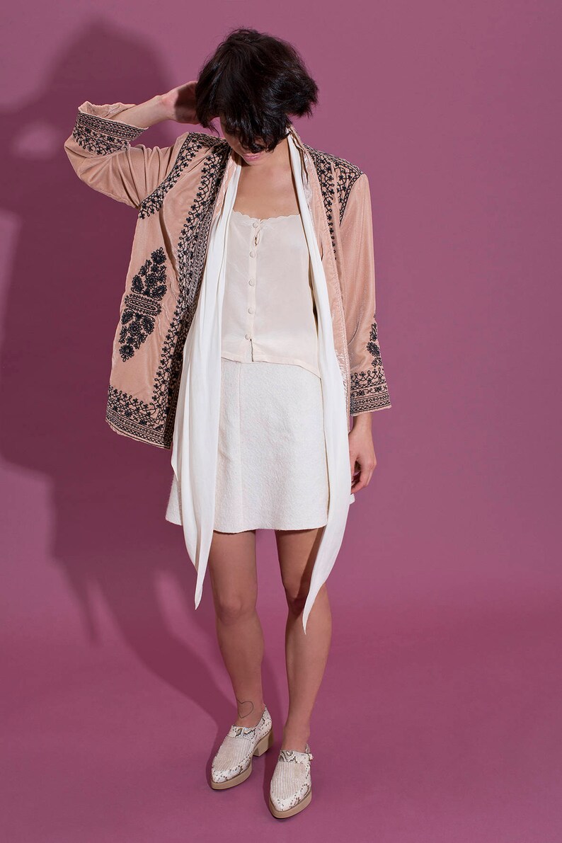 Puede incluir: Una mujer lleva una chaqueta rosa bordada con una bufanda blanca y una falda blanca. Lleva zapatos blancos con estampado de piel de serpiente.
