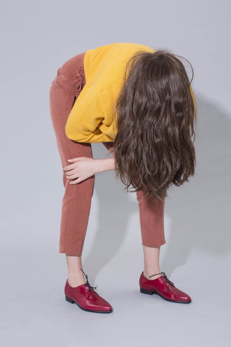 May include: A person wearing a yellow sweater and brown pants with red leather shoes. The person is bending over with their hair covering their face.