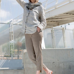 May include: A woman wearing a light gray linen shirt with a gray and white scarf, brown plaid pants, and brown leather shoes. She is standing on a concrete surface with a white bridge in the background.