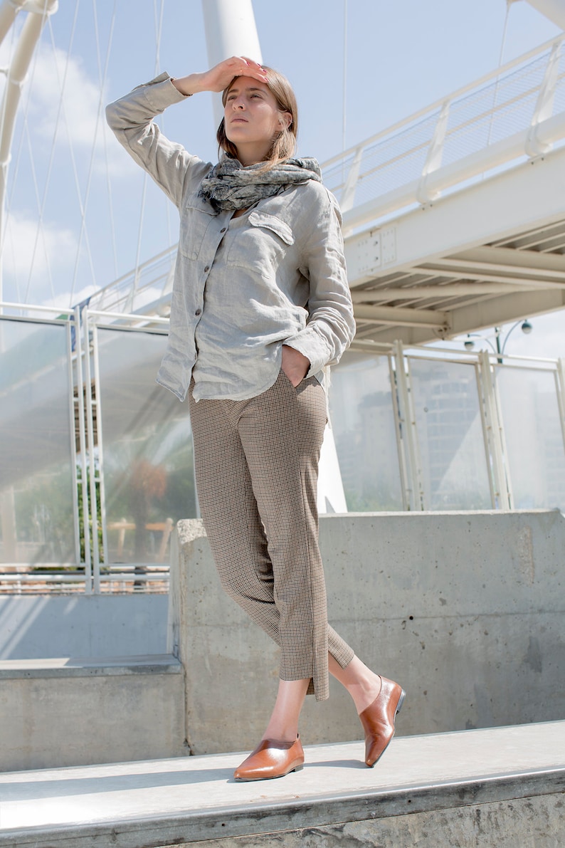 May include: A woman wearing a light gray linen shirt with a gray and white scarf, brown plaid pants, and brown leather shoes. She is standing on a concrete surface with a white bridge in the background.