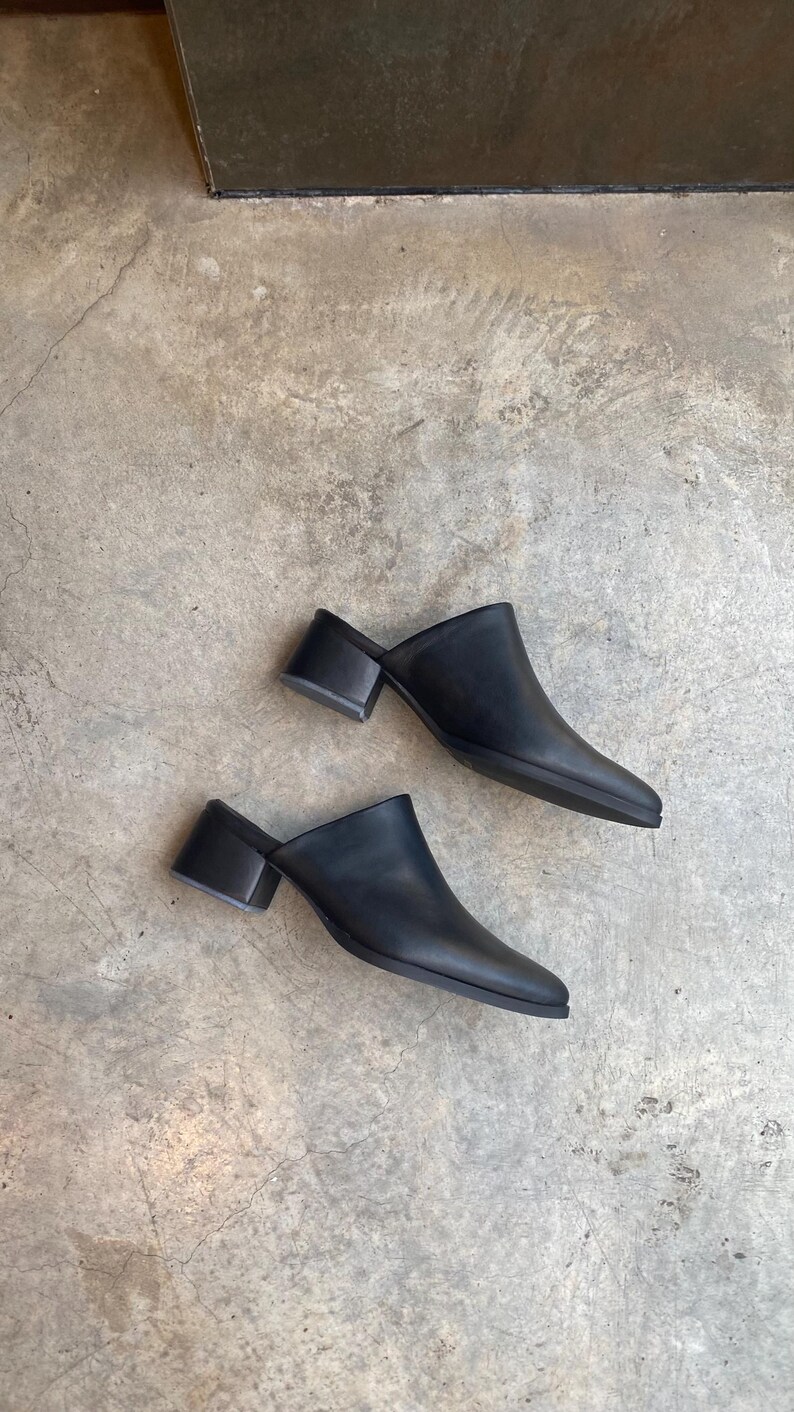 May include: A pair of black leather heeled mules with a pointed toe. The shoes are on a concrete surface.