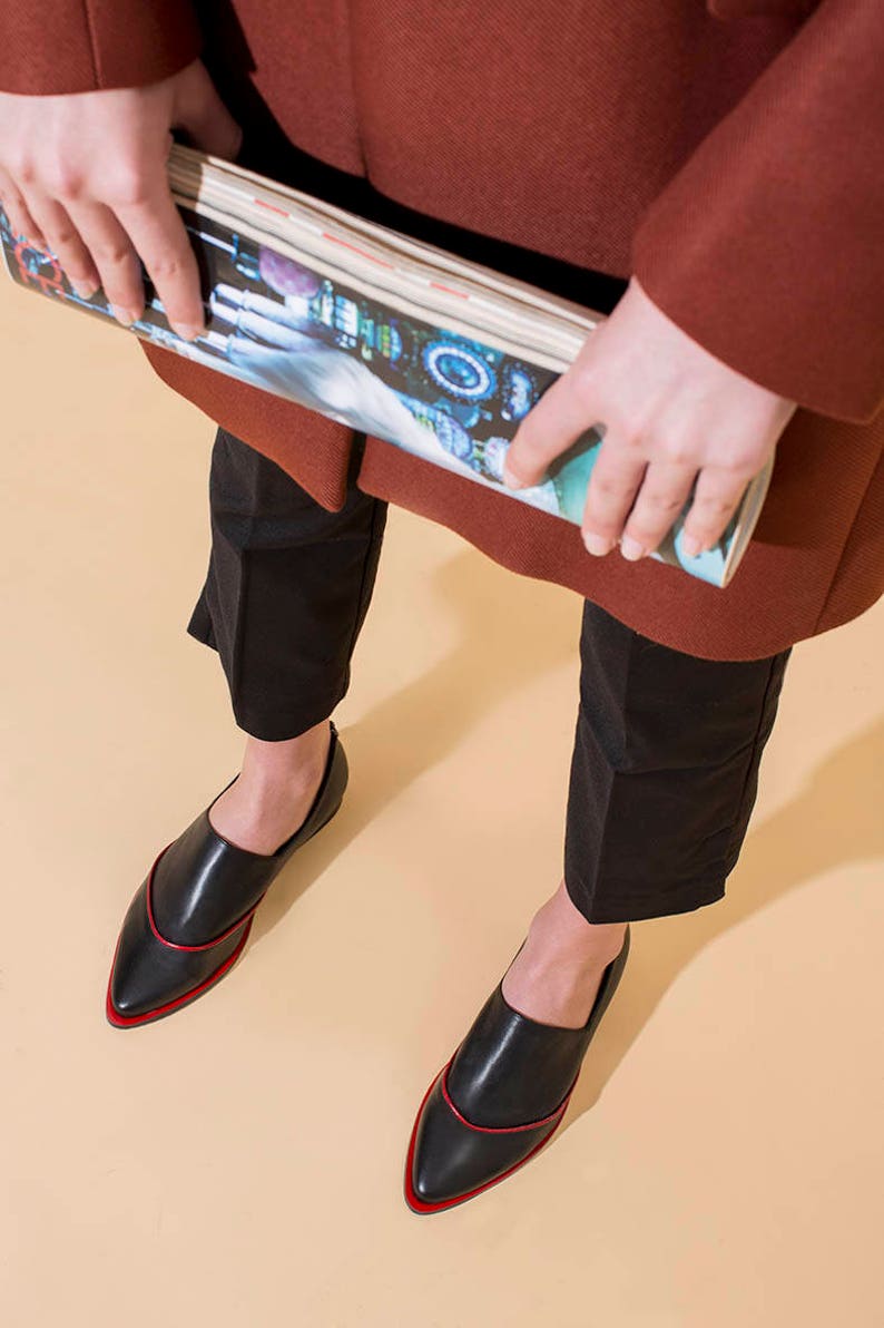 May include: Black leather shoes with a pointed toe and a red trim. The shoes are worn by a person wearing black pants and a brown coat.