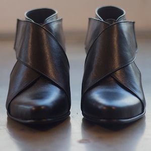 May include: A pair of black leather ankle boots. The boots have a distinctive design with straps that cross over the front. They have a pointed toe and a low heel. The leather has a subtle sheen, and the boots are set against a neutral background.