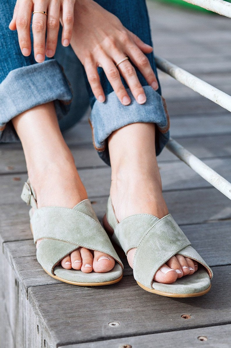 K&ouml;nnte beinhalten: Ein Paar hellgr&uuml;ne Wildledersandalen mit Kreuzriemen und einem Kn&ouml;chelriemen. Die Sandalen werden von einer Person getragen, die auf einer Holzoberfl&auml;che sitzt.