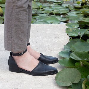 May include: A pair of black leather ankle strap shoes with pointed toes. The shoes are worn by a person standing on a stone path next to a pond with lily pads.