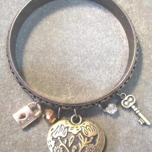 May include: A silver-coloured bangle bracelet with decorative charms. Charms include a heart with floral design, a padlock, a key, and a faceted bead. The bracelet has an etched pattern around the band.