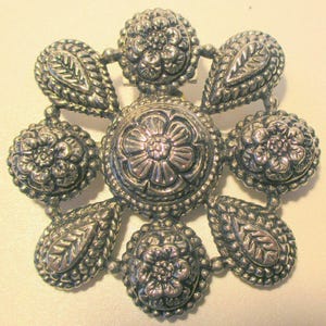 May include: A silver-toned brooch with a floral design. The brooch features a central flower surrounded by six smaller floral elements and six teardrop-shaped accents with leaf patterns. The metal has a detailed, textured finish.