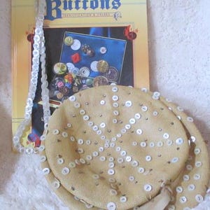 May include: A beige straw hat with a white button design. The hat has a white ribbon tied around it. The hat is sitting on a white fuzzy surface. A book titled "Antique & Collectible Buttons" is visible in the background.