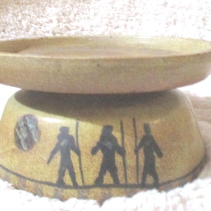 May include: A ceramic pedestal bowl with a round top and a base decorated with black silhouettes of figures. The bowl is a light tan color, with a darker tan base. The figures are holding staffs.