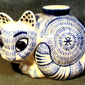 May include: A ceramic cat figurine featuring a cream-colored base with a blue hand-painted pattern of fine lines and dots. The cat has large black eyes, pointed ears with spotted details, and a circular opening on its back with a crackled glaze finish.