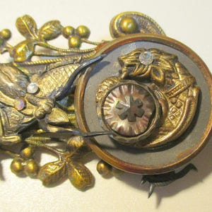 May include: Ornate antique metal brooch with a dragon and floral design. The brooch features gold and silver tones, with a central circular element depicting a lion and a starburst design. Embellished with rhinestones and iridescent accents.