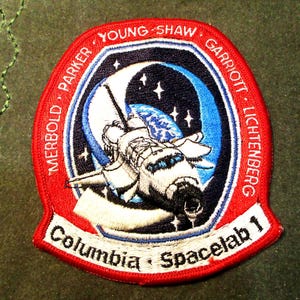 May include: Embroidered patch featuring a space shuttle in front of a blue and white planet and stars. The patch has a red border with the names of the crew members and the text "Columbia Spacelab 1".