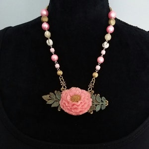 May include: A pink flower pendant necklace with gold and green accents. The necklace is made of pink and white beads.