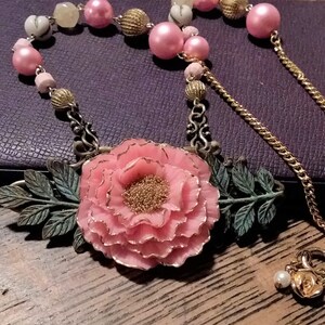 May include: A pink flower pendant necklace with gold chain and pink and white beads. The pendant features a large pink flower with gold accents and green leaves.