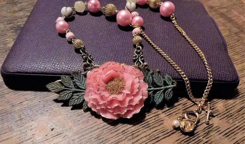 May include: A pink flower pendant necklace with gold chain and pink and white beads. The pendant features a large pink flower with gold accents and green leaves.