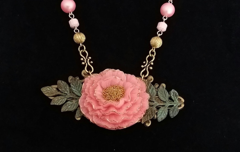 May include: A pink flower necklace with a gold chain and pink and white beads. The flower is surrounded by green leaves and has a gold center.