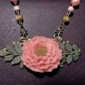 May include: A delicate pink flower pendant necklace with bronze chain and leaf accents. The flower is made of polymer clay and has a textured surface.