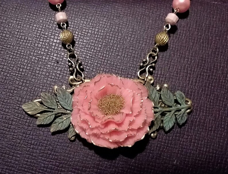 May include: A delicate pink flower pendant necklace with bronze chain and leaf accents. The flower is made of polymer clay and has a textured surface.
