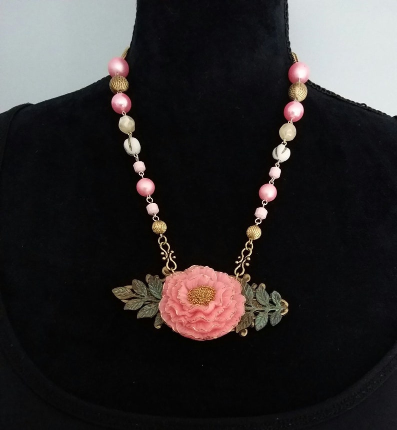 May include: A pink flower pendant necklace with gold and green accents. The necklace is made of pink and white beads.