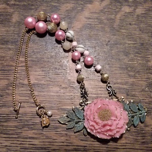 May include: A pink and gold necklace with a large pink flower pendant. The necklace features a chain with a clasp and a variety of beads, including pink, gold, and white.