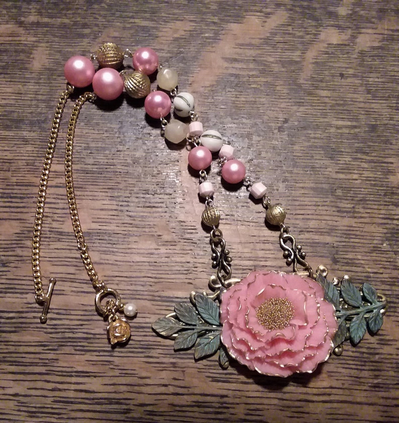 May include: A pink and gold necklace with a large pink flower pendant. The necklace features a chain with a clasp and a variety of beads, including pink, gold, and white.