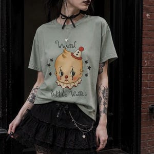 Peut inclure: Un t-shirt à manches courtes vert sauge avec une illustration d'un bébé clown triste portant un nez rouge et un chapeau de fête. Le texte "Weird little Wares" est imprimé en écriture noire au-dessus et en dessous, entouré de petites étoiles.