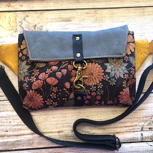 May include: A black and brown floral patterned fanny pack with a gray suede flap and a gold metal clasp. The bag has a black adjustable strap and a gold metal buckle. The bag has the text "Smoke & Meadow Leather" on the bottom right corner.