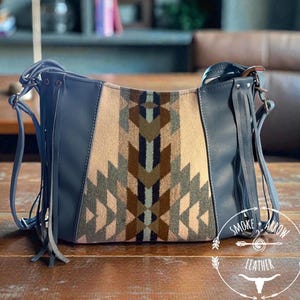 May include: A navy blue leather shoulder bag with a geometric patterned center panel in shades of tan, brown, and gray. The bag features fringe detailing and adjustable straps. The bag has the text "Smoke + Arrow Leather" on the lower right.