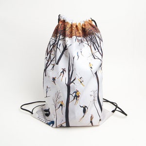 Peut inclure: Un sac à dos à cordon blanc orné d'une illustration répétée de personnes faisant du ski à travers une forêt d'arbres sombres et dénudés. Le haut du sac présente une bande de feuillage automnal orange-brun et des cordons noirs.