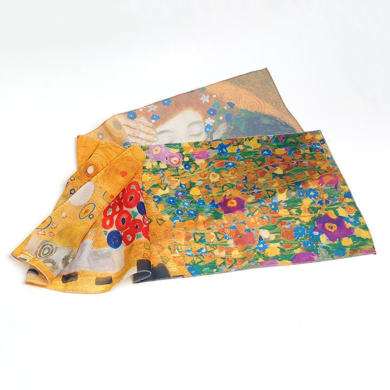 May include: A yellow and gold silk scarf with a colorful floral pattern and a portrait of a woman with red hair. The scarf is folded and draped on a white surface.