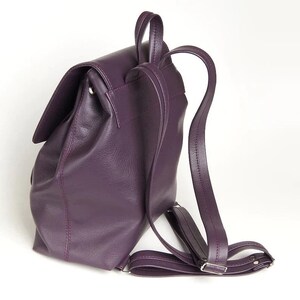 May include: A purple leather backpack with adjustable straps. The backpack has a flap closure and a single zippered pocket on the front.