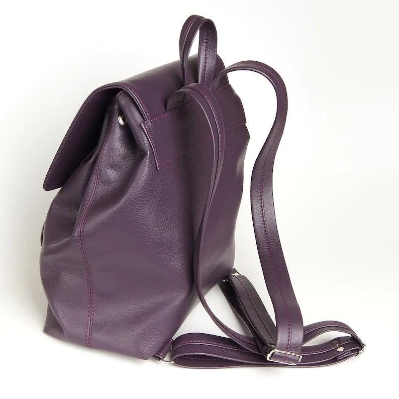 May include: A purple leather backpack with adjustable straps. The backpack has a flap closure and a single zippered pocket on the front.