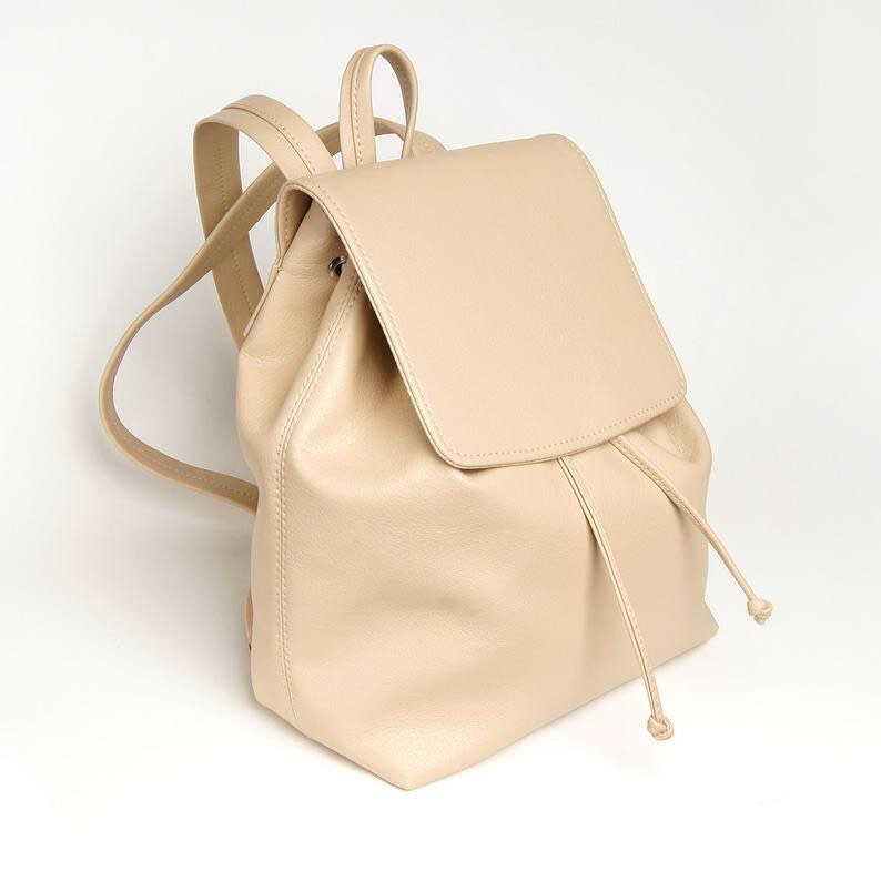 May include: A beige leather backpack with a flap closure and drawstring closure. The backpack has adjustable straps and a small pocket on the front.