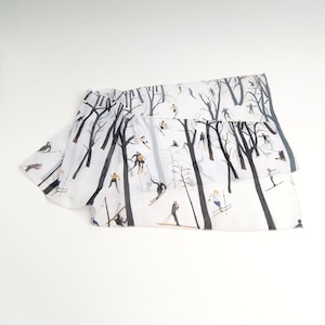 May include: White silk scarf with a black and white print of skiers in a snowy forest. The skiers are wearing colorful clothing and are skiing down a hill.