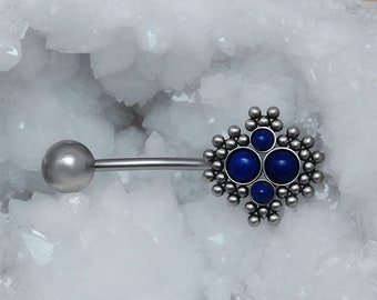 May include: A silver-colored body piercing barbell with a decorative end featuring four deep blue gemstones surrounded by small silver beads. The barbell is placed on a white, crystalline surface.
