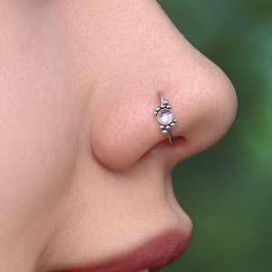 以下が含まれることがあります： ピンクの宝石が付いた小さな銀製の鼻のリングをした人の鼻のクローズアップ。リングは宝石の周りに小さな装飾ビーズが付いています。