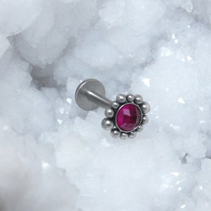 Puede incluir: Un piercing de cuerpo plateado con una pequeña gema redonda rosa en el centro rodeada por un halo plateado de pequeñas cuentas.