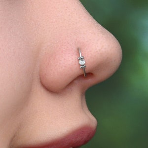May include: A silver nose ring with a small, clear gemstone and two decorative beads. The nose ring is worn on the left nostril. The background is a blurred green.
