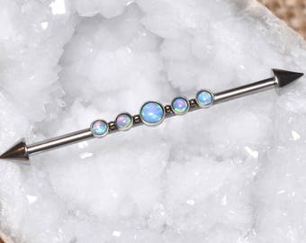 Puede incluir: Una joya corporal de barra industrial recta con un eje de tono plateado metálico y extremos en forma de cono puntiagudo. El centro de la barra está adornado con cinco piedras redondas de color azul claro con aspecto de ópalo iridiscente.