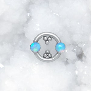 May include: A circular silver-toned body jewelry ring featuring two round, iridescent blue opal cabochons set on opposite sides. The inner circumference of the ring is accented with two clusters of three small, polished silver-toned metal spheres.